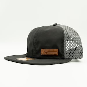 Può includere: Un cappello da baseball in mesh nero e grigio con una toppa in pelle marrone che presenta un'icona di bicicletta. Il cappello ha una visiera piatta e una parte posteriore in mesh.
