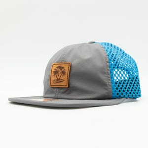 May include: A gray and blue mesh baseball cap with a brown leather patch featuring a palm tree design.
