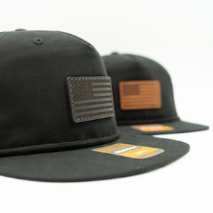 Könnte beinhalten: Zwei schwarze Baseballkappen mit flachem Schirm und einem braunen Lederaufnäher mit der amerikanischen Flagge. Die Kappen haben eine strukturierte Passform und ein Seildetail am Schirm. Auf weißem Hintergrund.