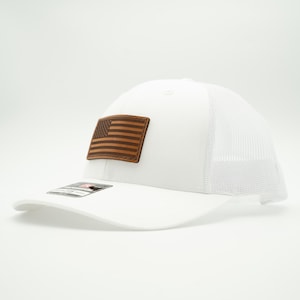 May include: White trucker hat with a brown leather patch featuring an American flag design. The hat has a curved brim and a mesh back for breathability. A classic and patriotic accessory.
