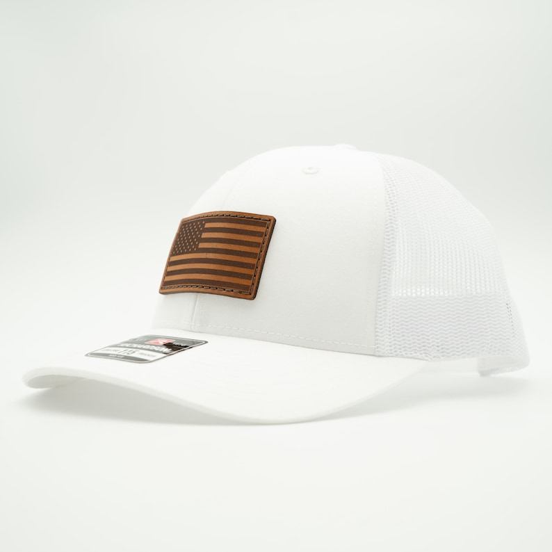 May include: White trucker hat with a brown leather patch featuring an American flag design. The hat has a curved brim and a mesh back for breathability. A classic and patriotic accessory.