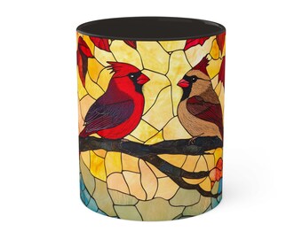 May include: A stained glass style mug featuring two red cardinals perched on a branch. The background is a yellow and orange stained glass design.