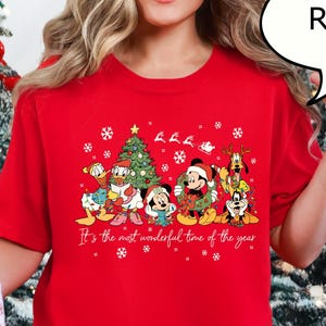 May include: A red Christmas t-shirt featuring Disney characters, including Mickey Mouse, Donald Duck, and Goofy, with a Christmas tree and the text "It's the most wonderful time of the year." The shirt is paired with red and black plaid pajama pants.