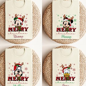 Op de afbeelding: Zes witte T-shirts met een rood-zwart ruitpatroon aan de bovenkant. Elk shirt heeft een ander Disney-personage dat een kerstmanmuts en rendiergewei draagt. De tekst "Merry Christmas" staat op elk shirt gedrukt met een naam eronder. De namen zijn "Daddy", "Mommy", "Big Sis", "Lil Bro", "Also" en "Jacie".
