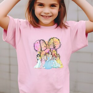 Puede incluir: Una camiseta rosa claro con un gr&aacute;fico colorido de un lazo con orejas de rat&oacute;n y varias princesas frente a un castillo. El gr&aacute;fico est&aacute; centrado en la parte delantera de la camiseta.