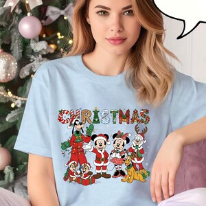 May include: Pale blue t-shirt featuring the word "CHRISTMAS" in colourful letters and cartoon characters. The shirt shows Mickey Mouse, Minnie Mouse, Goofy, and other Disney characters in festive outfits. The word "CHAMBRAY" is in a speech bubble.
