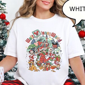 May include: White t-shirt with a colorful graphic featuring Disney characters in a Christmas theme. The text "Walt Disney World" is displayed above the characters. The shirt is part of a holiday collection.
