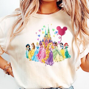 May include: A white t-shirt with a colorful graphic of Disney princesses standing in front of Cinderella's castle. The graphic also includes a pink Mickey Mouse balloon with hearts floating around the castle.