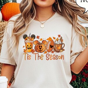 May include: Ivory-colored t-shirt featuring a fall-themed design. The design includes a pumpkin spice latte with a Mickey Mouse face, a Mickey Mouse pumpkin, a football with a Mickey Mouse face, and a mug of hot cocoa. The words 'Tis The Season' are printed in orange.