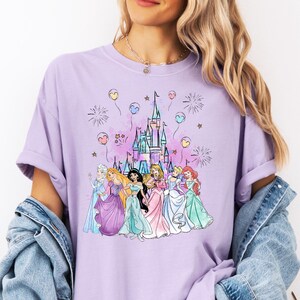 Puede incluir: Una camiseta morada con un gr&aacute;fico del castillo de Disney y siete princesas. Las princesas llevan sus vestidos caracter&iacute;sticos y caminan hacia el castillo. Hay globos y fuegos artificiales en el fondo.