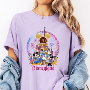 May include: A purple t-shirt with a graphic of Mickey Mouse and friends celebrating Disneyland's 70th anniversary. The graphic features the Disneyland castle, fireworks, and the text "Disneyland Resort".
