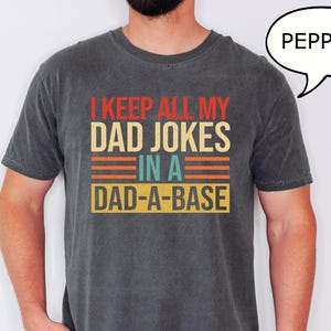 May include: A dark gray t-shirt with the text "I KEEP ALL MY DAD JOKES IN A DAD-A-BASE" in red, orange, and yellow. The shirt is a crew neck and has short sleeves. The person is wearing a watch with a red band.