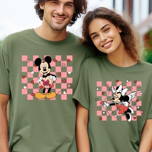 Puede incluir: Camisetas verde oliva con dise&ntilde;os de Mickey y Minnie Mouse. Mickey sostiene un regalo en forma de coraz&oacute;n sobre un fondo a cuadros rosa y blanco. Minnie est&aacute; vestida como Cupido con arco y flecha.