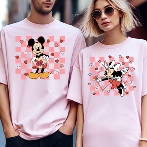 Puede incluir: Camisetas rosas con dise&ntilde;os de Mickey y Minnie Mouse. Una camiseta tiene a Mickey sosteniendo un regalo en forma de coraz&oacute;n, la otra tiene a Minnie como Cupido con arco y flecha. Ambas camisetas tienen un fondo a cuadros rosa y blanco con corazones.
