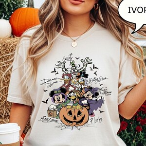 May include: Ivory-colored t-shirt featuring a Halloween-themed graphic with cartoon characters, including Mickey Mouse, Donald Duck, and Goofy. The design includes a carved pumpkin and bats. The shirt is worn by a person in blue jeans.
