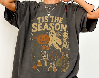 Może przedstawiać: Ciemnoszara koszulka oversize z motywem Halloween. Projekt zawiera dynię, ducha, szkielet i napis "Tis the Season". Inne elementy to cukierki, butelki z miksturami i nagrobek.