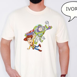 May include: Ivory-colored t-shirt featuring a graphic of Woody and Buzz Lightyear from Toy Story. Woody is wearing a cowboy hat and Buzz is in a spacesuit. The word "IVORY" is in a speech bubble.