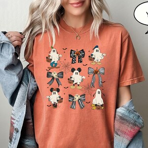 Puede incluir: Camiseta naranja con un dise&ntilde;o tem&aacute;tico de Halloween. El dise&ntilde;o incluye fantasmas de dibujos animados con lazos, calabazas y telara&ntilde;as. La palabra "YAM" est&aacute; en un bocadillo. La camiseta se usa con una chaqueta vaquera.