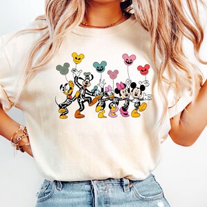 May include: A white t-shirt with a graphic of Mickey Mouse, Minnie Mouse, Donald Duck, Goofy, and Pluto dressed in skeleton costumes holding colorful balloons. The balloons are shaped like Mickey Mouse ears and have a Halloween theme.