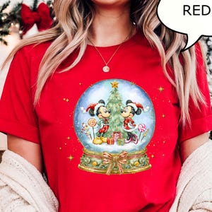 May include: A red t-shirt featuring a snow globe design with Mickey and Minnie Mouse in holiday attire. The snow globe includes a Christmas tree, gifts, and candy. The word "RED" is in a speech bubble.