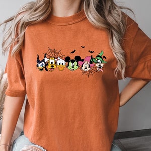 May include: Orange t-shirt with a Halloween-themed graphic featuring Mickey Mouse, Minnie Mouse, Donald Duck, Goofy, and other Disney characters. The characters are wearing costumes and are surrounded by bats and a spider web.
