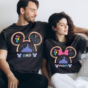 Pode incluir: Um casal vestindo camisetas pretas combinando com um design de silhueta do Mickey Mouse. As camisetas apresentam uma imagem de desenho animado do castelo da Cinderela, bal&otilde;es e fogos de artif&iacute;cio. A camiseta do homem diz "DAD" e a camiseta da mulher diz "MOM".