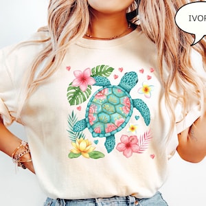 May include: Ivory-colored t-shirt featuring a turquoise sea turtle with a floral design on its shell. The turtle is surrounded by pink and yellow flowers, green leaves, and small pink hearts. The word "IVORY" is in a speech bubble.