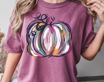 May include: A mauve t-shirt with a colorful abstract pumpkin design. The pumpkin is outlined in black and has a variety of colors including pink, blue, yellow, and orange.