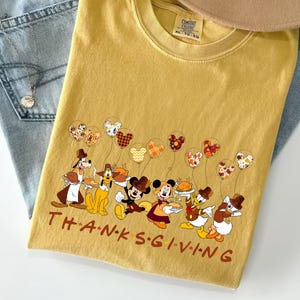 May include: Mustard-colored t-shirt with a Thanksgiving-themed graphic featuring cartoon characters and balloons. The text "THANKSGIVING" is printed across the bottom. A tan hat and blue jeans are also in the image.