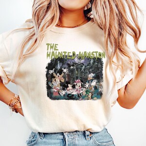 May include: A white t-shirt with a vintage-style graphic of Mickey Mouse and friends in front of the Haunted Mansion. The text "THE HAUNTED MANSION" is printed in green above the graphic.