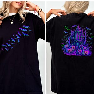 May include: Black t-shirt featuring a Halloween theme. The front displays a flock of bats in shades of blue and purple. The back showcases a haunted house, pumpkins, and bats in vibrant neon colors. A white collar is visible.