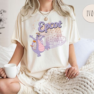 May include: A cream colored t-shirt with a purple dragon and the text "Epcot" in a gold font. The dragon is flying in front of a stylized image of the Spaceship Earth attraction at Epcot.