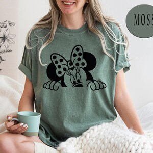 K&ouml;nnte beinhalten: Ein gr&uuml;nes T-Shirt mit einer schwarzen und wei&szlig;en Grafik von Minnie Mouse, die &uuml;ber den Rand schaut. Der Text "MOSS" befindet sich in einem gr&uuml;nen Kreis in der oberen rechten Ecke des Bildes.