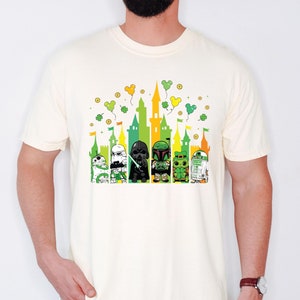 Puede incluir: Camiseta color crema con un gr&aacute;fico colorido de personajes de Star Wars frente a una silueta de castillo. El dise&ntilde;o incorpora tonos verdes, amarillos y naranjas con tr&eacute;boles y globos de Mickey Mouse, celebrando el D&iacute;a de San Patricio.