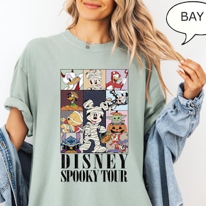 May include: A light green t-shirt featuring a grid of Disney characters in Halloween costumes. The shirt has the words "DISNEY SPOOKY TOUR" printed in black. The shirt is a relaxed fit.