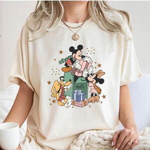 May include: A cream coloured t-shirt featuring a vintage cartoon graphic of Mickey Mouse, Minnie Mouse, and Pluto standing by a mailbox with presents and stars. The text "Mail" is on the mailbox.