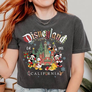 Puede incluir: Una camiseta negra con un logotipo de Disneyland de estilo vintage, con Mickey Mouse, Donald Duck, Goofy y Pluto frente al Castillo de la Bella Durmiente. El texto dice "Disneyland Est. 1955 California".