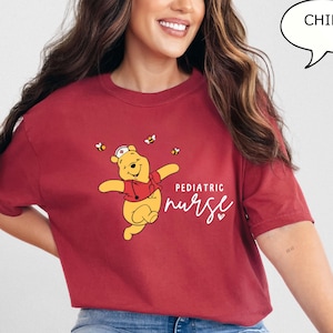 May include: A maroon t-shirt featuring a cartoon bear wearing a nurse's hat and stethoscope, with the words "Pediatric Nurse" in white script. The design includes small bees. The shirt is worn by a person.