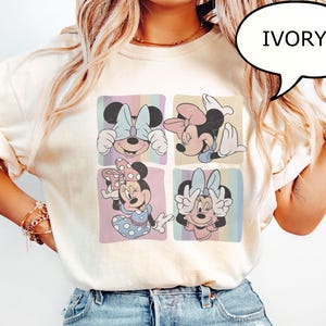 Puede incluir: Camiseta de color marfil con un gr&aacute;fico de cuatro paneles de Minnie Mouse en varias poses. Cada panel tiene un fondo de rayas pastel. La palabra "IVORY" est&aacute; en una burbuja de di&aacute;logo. La camiseta se combina con vaqueros azules.