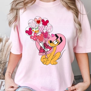 Może przedstawiać: R&oacute;żowy T-shirt z kresk&oacute;wkowym psem trzymającym pudełko czekoladek w kształcie serca. Pies jest otoczony r&oacute;żowymi i czerwonymi balonami w kształcie serca.