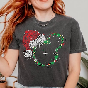 Puede incluir: Camiseta gris oscuro con un dise&ntilde;o navide&ntilde;o. El gr&aacute;fico incluye un gorro de Pap&aacute; Noel y una corona con forma de Mickey Mouse, ambos compuestos por puntos de colores. El dise&ntilde;o incorpora elementos rojos, blancos, verdes y amarillos, creando un tema festivo.