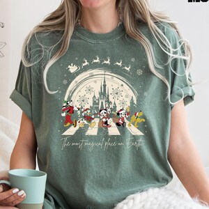 Puede incluir: Camiseta verde con una ilustraci&oacute;n de estilo vintage de Mickey Mouse y sus amigos caminando por un paso de cebra frente al Castillo de Cenicienta. El texto "The most magical place on Earth" est&aacute; impreso debajo de la ilustraci&oacute;n.