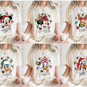 May include: Six cream-colored t-shirts featuring Disney characters in festive Christmas designs. Each shirt showcases a different character: Mickey Mouse, Minnie Mouse, Pluto, Donald Duck, Daisy Duck, and Goofy, all wearing holiday attire. Each shirt has the character's name.