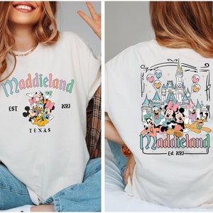 K&ouml;nnte beinhalten: Ein wei&szlig;es T-Shirt mit einem bunten Motiv von Mickey Mouse und seinen Freunden vor einem Schloss. Der Text "Maddieland Est. 2023" ist auf dem Shirt aufgedruckt.