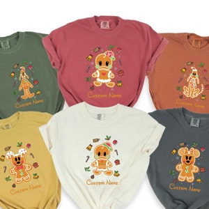 May include: Six different colored t-shirts with a gingerbread character design. Each shirt has a different character, including Mickey Mouse, Donald Duck, and Pluto. The shirts are all in festive holiday colors and have the text "Custom Name" printed on the bottom.