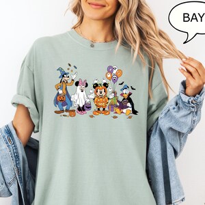 May include: A light green t-shirt featuring a Halloween-themed graphic of Disney characters, including Mickey Mouse, Minnie Mouse, Donald Duck, and Goofy, dressed in costumes. The word "BAY" is in a speech bubble.