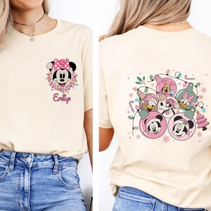 K&ouml;nnte beinhalten: Cremefarbenes T-Shirt mit einem Minnie Mouse-Motiv mit rosa Schleife und Blumenkranz sowie dem Namen "Emily". Die R&uuml;ckseite des Shirts zeigt Disney-Figuren in Weihnachtsbaumschmuck-Designs.