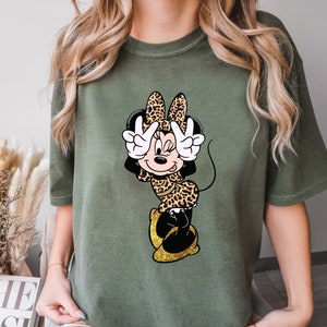 K&ouml;nnte beinhalten: Olivgr&uuml;nes T-Shirt mit einer Cartoon-Minnie-Maus in einem Leopardenmuster-Outfit. Minnie macht mit beiden H&auml;nden ein Friedenszeichen. Ihre Schuhe sind goldfarben glitzernd. Das Shirt ist ein l&auml;ssiger, bequemer Stil.