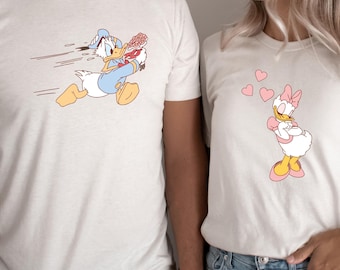May include: A matching pair of t-shirts featuring cartoon characters. The man's shirt shows Donald Duck running with a bouquet of flowers. The woman's shirt shows Daisy Duck with hearts floating above her head.