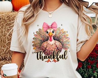 Könnte beinhalten: Elfenbeinfarbenes T-Shirt mit einem farbenfrohen Truthahn-Motiv, einer rosa Schleife und dem Wort "Thankful". Das Design enthält Glitzerakzente. Der Hintergrund zeigt Kürbisse und Heuballen, ideal für Thanksgiving.